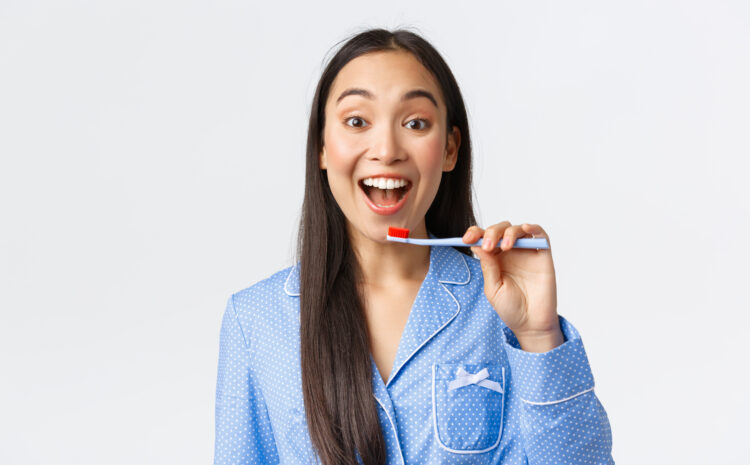 Cheerful pretty young asian girl in blue pajama waking-up, brushing teeth with broad enthusiastic smile, holding toothbrush near white teeth, white background. Copy space